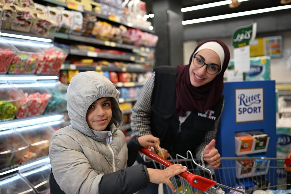A smiling boy pushes a shopping cart with an aid worker