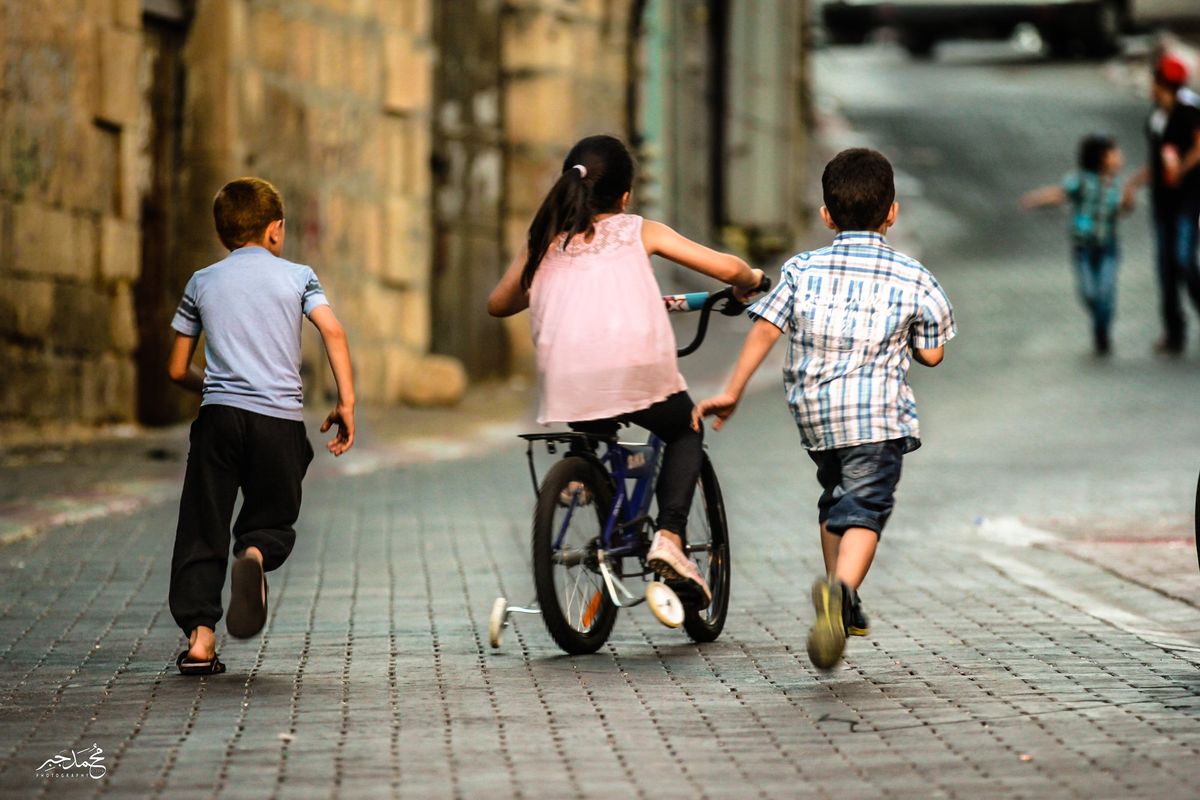 Children running beside a girl on a bike in an old-city lane